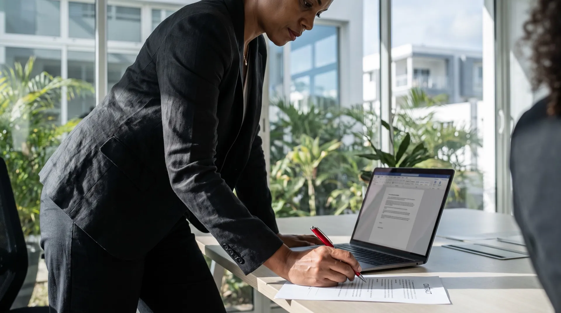 Dans un espace de travail lumineux, une dirigeante annote au stylo rouge un contrat imprimé, son ordinateur portable ouvert à côté