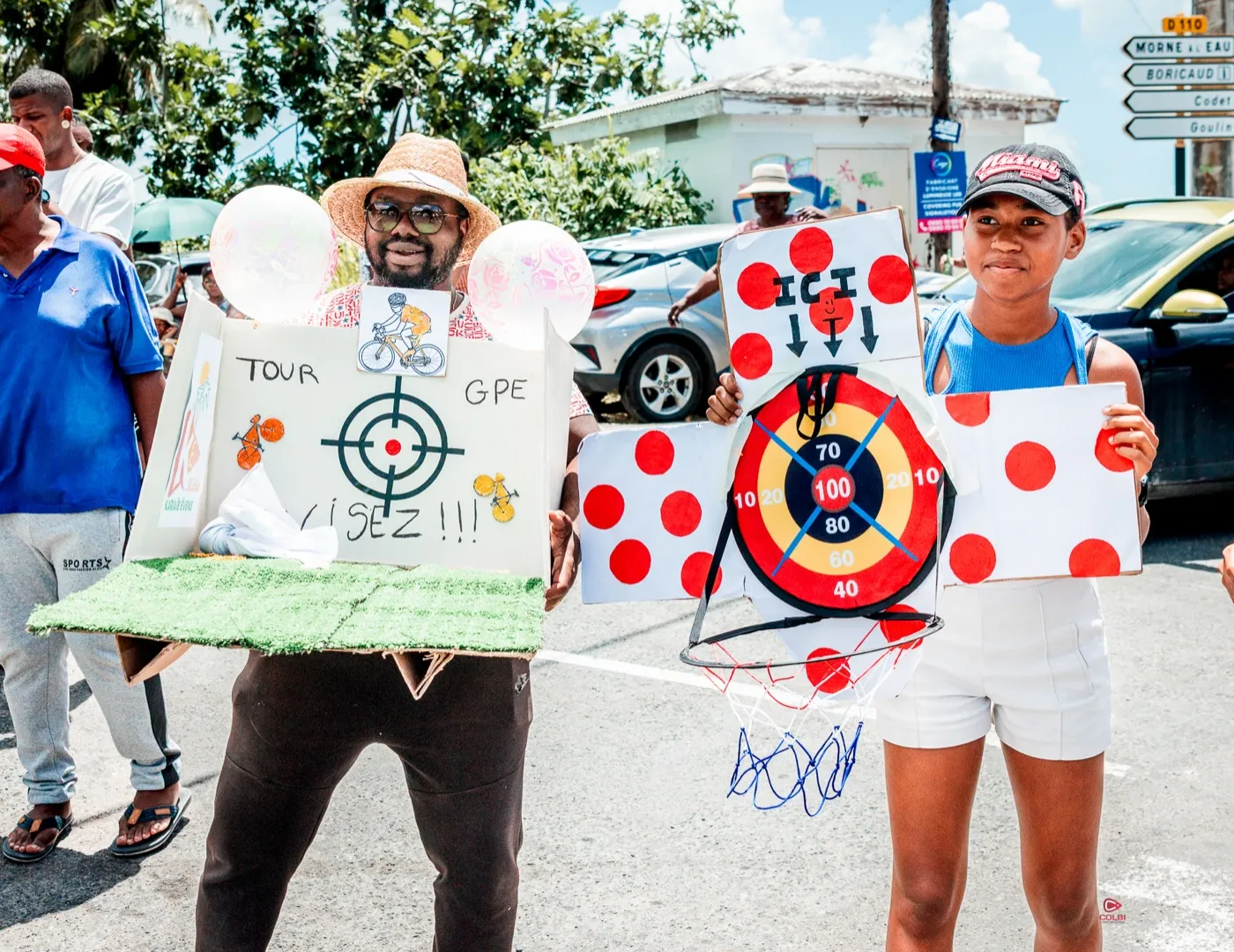 Spectateurs du Tour de Guadeloupe brandissant des cibles artisanales « Visez ! » et un panier de basket pour attraper les goodies de la caravane