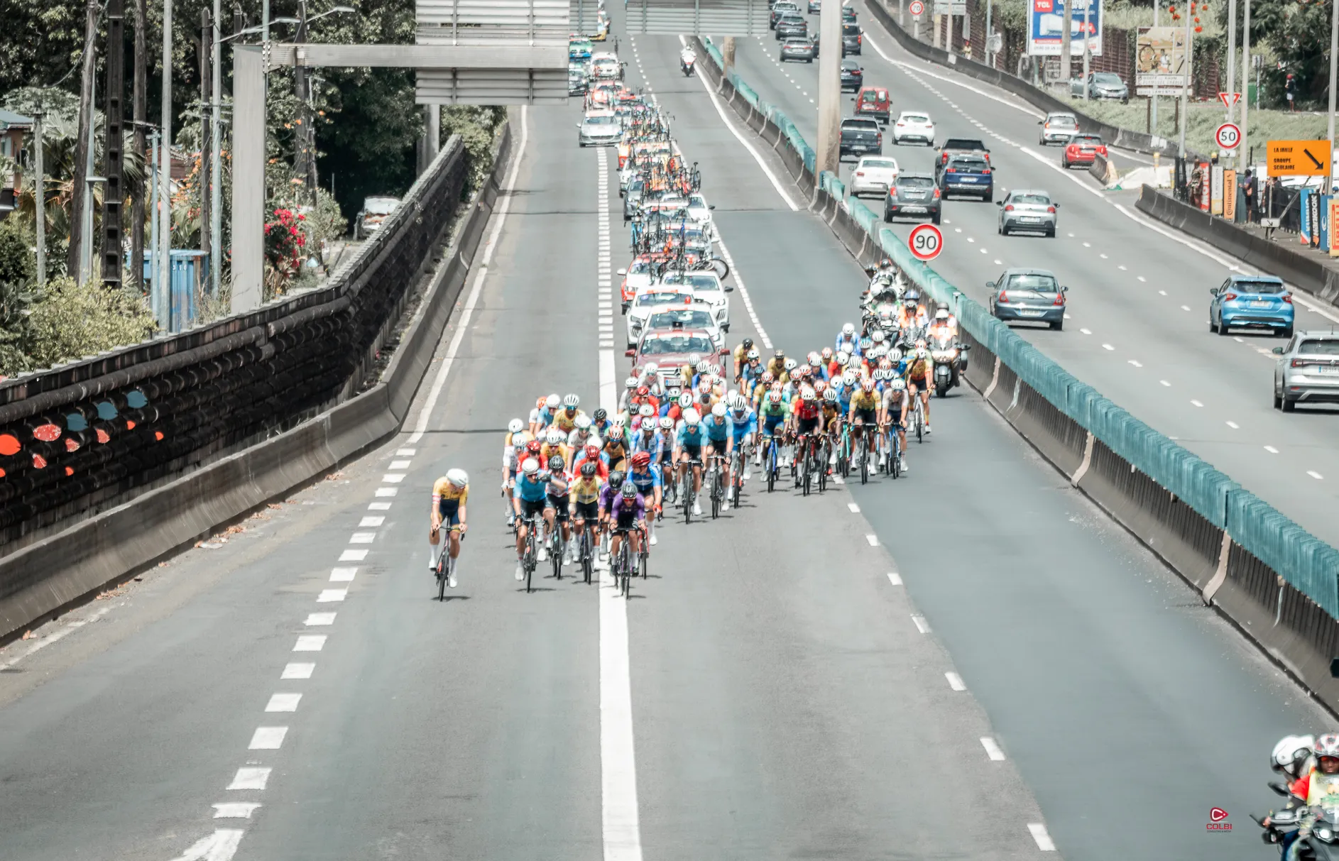 Le peloton du Tour cycliste de la Guadeloupe sur une voie rapide, vu d’une passerelle, suivi par les motos et les voitures de course.